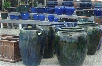 A selection of large ceramic pots in various shades of blue and green, displayed outdoors on a gravel surface. The pots vary in size and shape, with some featuring a 