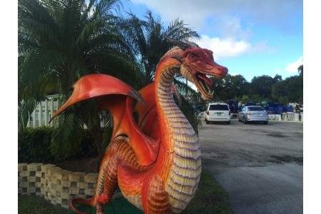 A large, red dragon sculpture with detailed scales stands in a parking lot next to a palm tree on a sunny day.